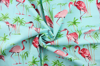 This outdoor fabric features a flamingo and tree design in green, pink, light brown, white and black against a light blue . 
