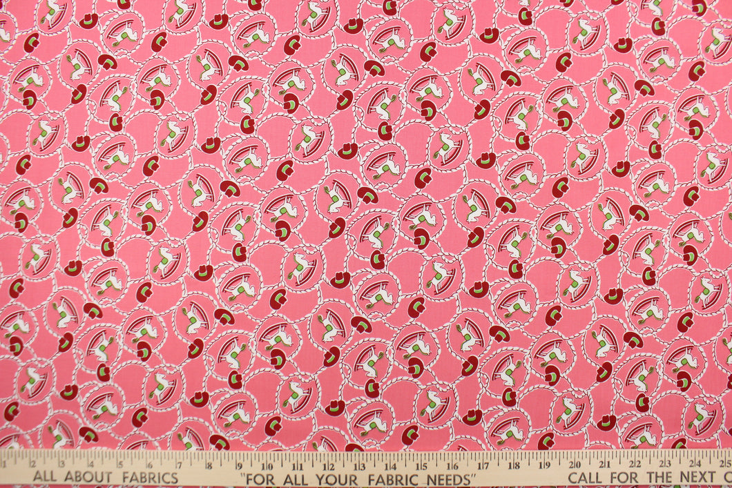 This quilting fabric features a cowboy/cowgirl theme with a rocking horse, rope and hat design in green, dark red, and white set against a medium pink background. 