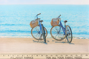 This fabric features a beautiful realistic view of two bikes along the seashore in beige, blue, black and gray.