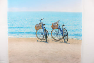 This fabric features a beautiful realistic view of two bikes along the seashore in beige, blue, black and gray.