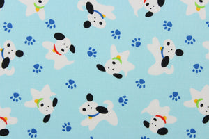 This cute quilting print features a white dog with black ears and different colored collars mix in with blue paw prints on a pale blue background. 