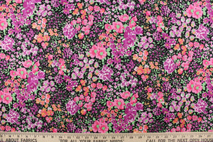 This quilting print features a floral design in varying shades of pink, purple, green and pale orange with hints of white against a black background. 